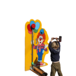 Ein Junge holt mit einem Hammer aus, um bei Hau den Lukas zu gewinnen. Dieser Hau den Lukas ist im Design eines Clowns, der Luftballons in der Hand hält.