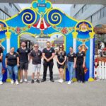 Eine Gruppe von Menschen steht vor einer blauen Eingangsfassade. Im Hintergrund ist ein Karussell von Ertl zu sehen.